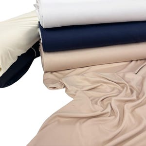May include: Several rolls of fabric in shades of white, navy, and beige. The textiles are neatly stacked and rolled, with some fabrics draped to show their texture. The image highlights the different colours and the smooth appearance of the materials.