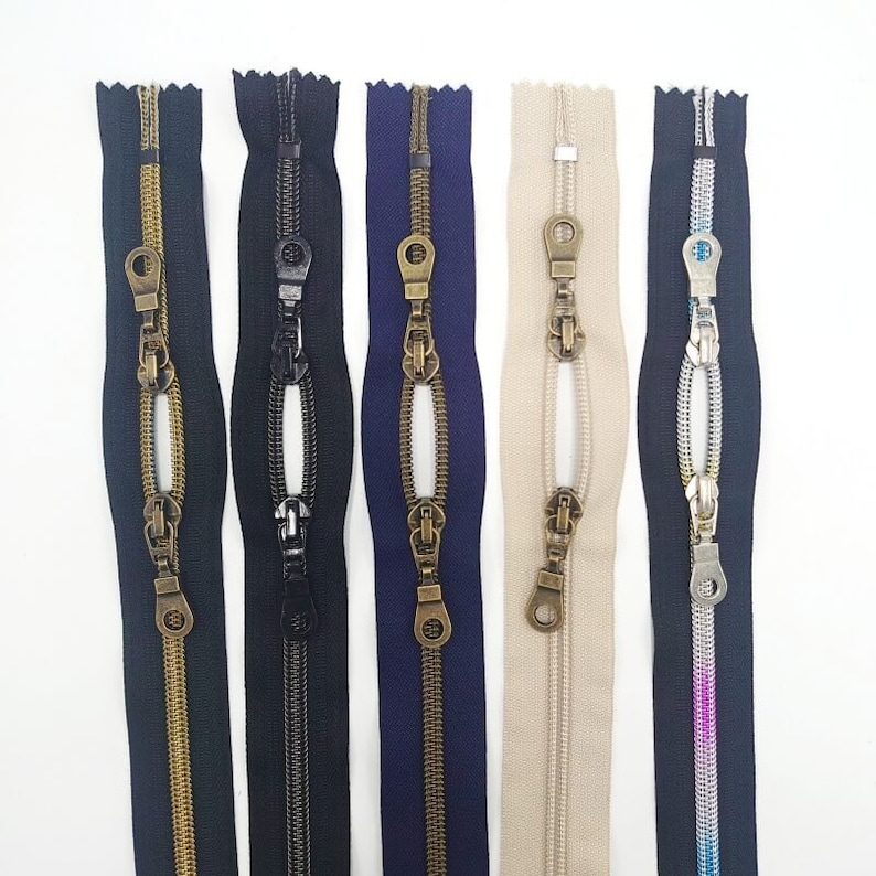 May include: Five different zipper pulls with metal sliders and teeth. The zippers are in black, navy blue, beige, silver, and pink.