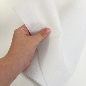 May include: White interfacing fabric, a type of fusible material used in sewing and crafting projects. The fabric is folded in half and held in a hand.