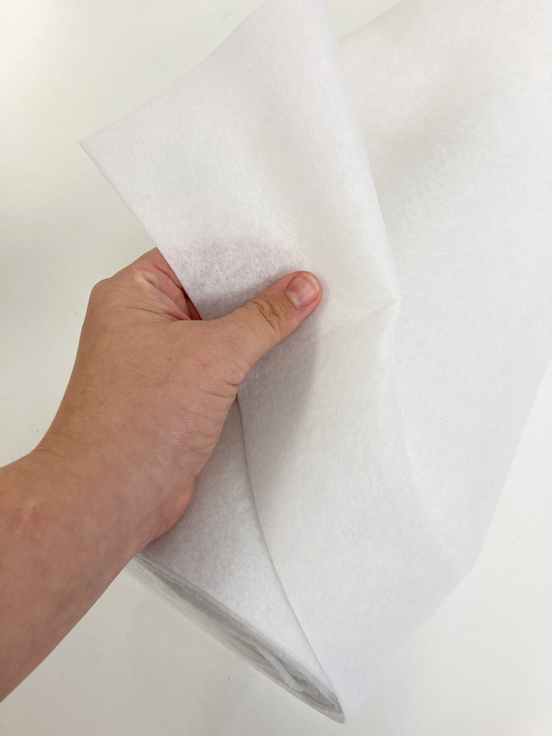 May include: White interfacing fabric, a type of fusible material used in sewing and crafting projects. The fabric is folded in half and held in a hand.