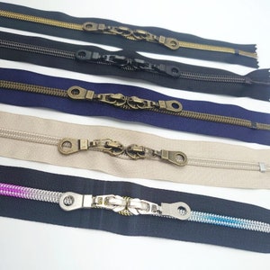 May include: Five different zipper pulls with various finishes. The pulls are made of metal and have a decorative design. The zippers are in different colors, including gold, black, blue, beige, and black with a silver pull.