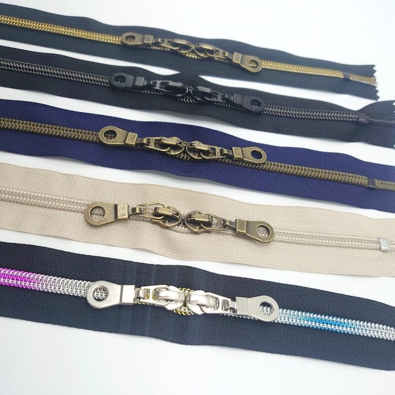 May include: Five different zipper pulls with various finishes. The pulls are made of metal and have a decorative design. The zippers are in different colors, including gold, black, blue, beige, and black with a silver pull.