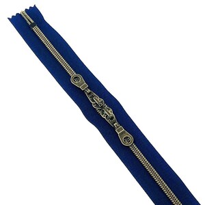 May include: A close-up of a blue zipper with a brass slider and pull. The zipper is closed and the slider is in the center of the zipper.
