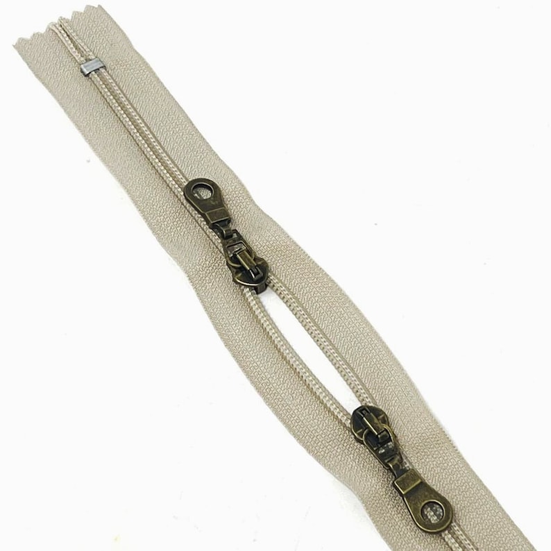 May include: A beige zipper with two antique brass sliders. The zipper is open and the sliders are visible.