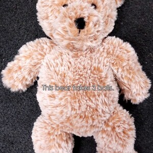 Pu&ograve; includere: Un orsacchiotto di peluche marrone chiaro con naso nero e occhi neri. L'orso &egrave; seduto su una superficie grigio scuro. Il testo "This bear takes 3 balls." &egrave; stampato sulla pancia dell'orso.