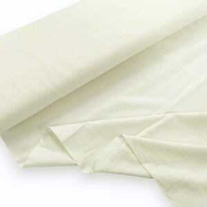May include: A large roll of solid off-white fabric featuring a smooth, matte texture. The material is draped in soft folds, showcasing a consistent light cream color throughout the entire length of the textile as it extends across the frame.