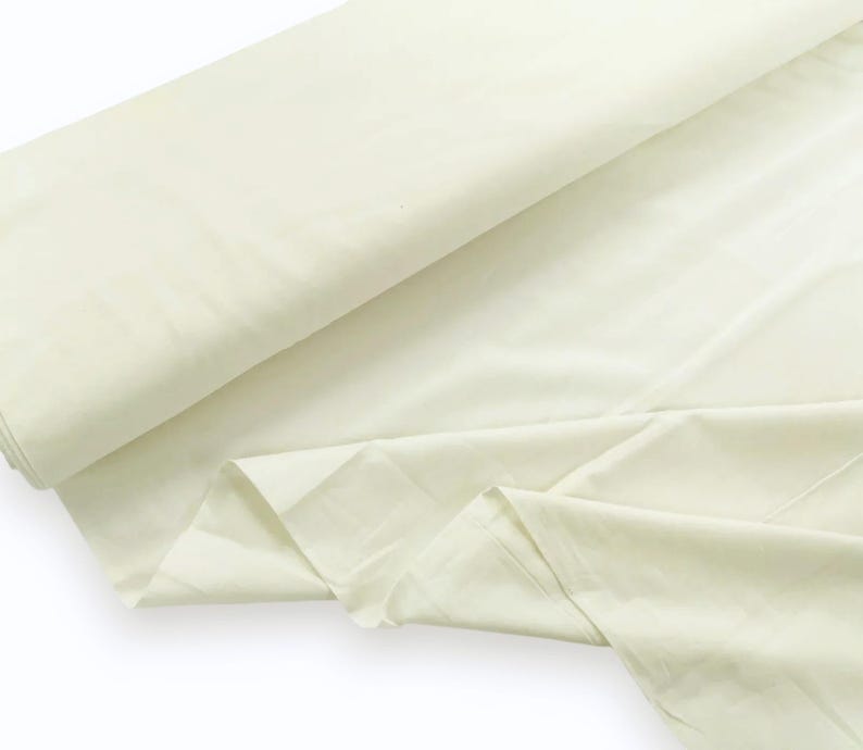May include: A large roll of solid off-white fabric featuring a smooth, matte texture. The material is draped in soft folds, showcasing a consistent light cream color throughout the entire length of the textile as it extends across the frame.