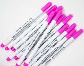 May include: A collection of white textile pens with bright pink caps and tips. Each pen has text that reads "Please test on fabric before use". The pens are designed for fabric marking and are water erasable.