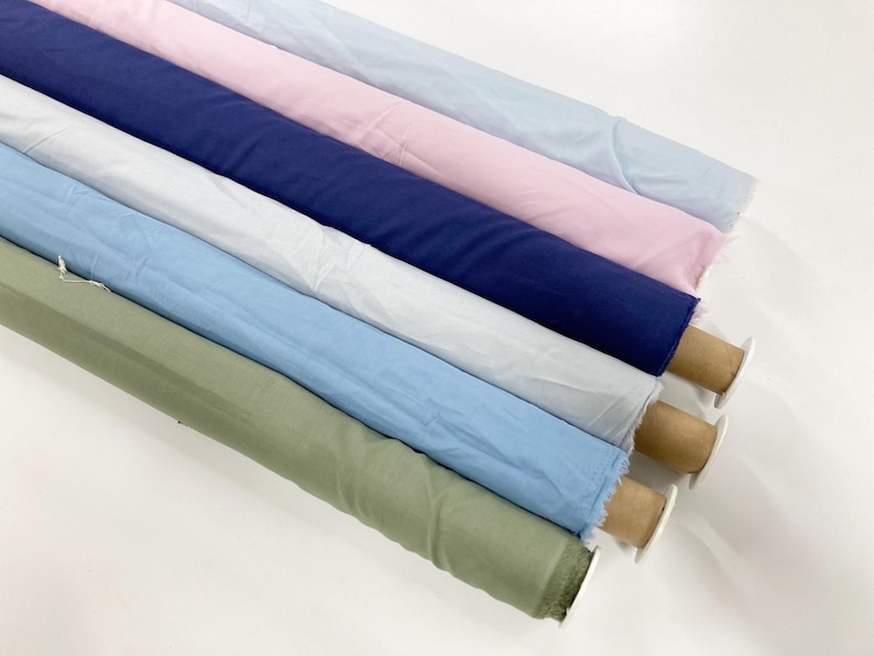 May include: Six rolls of fabric arranged diagonally, featuring solid colors including light blue, soft pink, navy blue, white, sky blue, and sage green. Each roll is wrapped around a cylindrical cardboard core, displaying a smooth texture.
