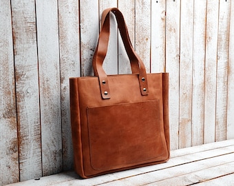 Pode incluir: Uma bolsa tote de couro marrom com um bolso frontal e duas alças. A bolsa está sobre uma superfície de madeira branca.