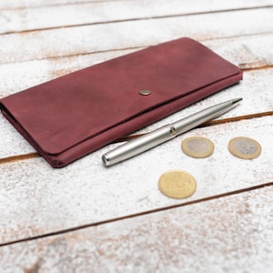 May include: A burgundy leather wallet with a silver pen and three silver coins on a white wooden surface.