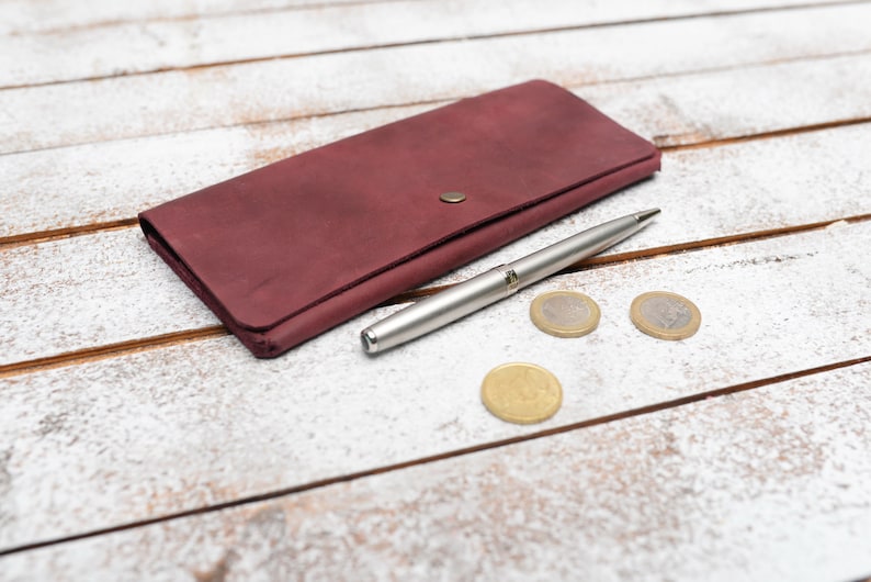 May include: A burgundy leather wallet with a silver pen and three silver coins on a white wooden surface.