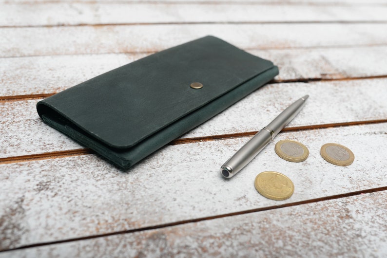 May include: A dark green leather wallet with a gold button closure, a silver pen, and three gold coins on a white wooden surface.