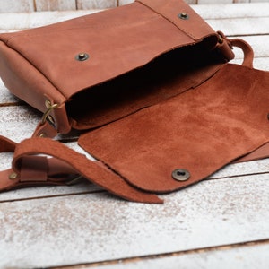 May include: A brown leather crossbody bag with a flap closure and a long strap. The bag is open and the inside is lined with brown suede.