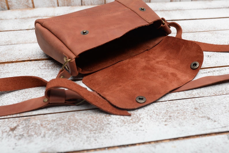 May include: A brown leather crossbody bag with a flap closure and a long strap. The bag is open and the inside is lined with brown suede.