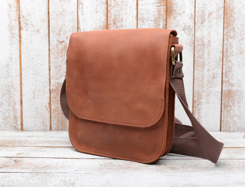 May include: A brown leather messenger bag with a brown adjustable strap. The bag has a flap closure and a single pocket on the front.