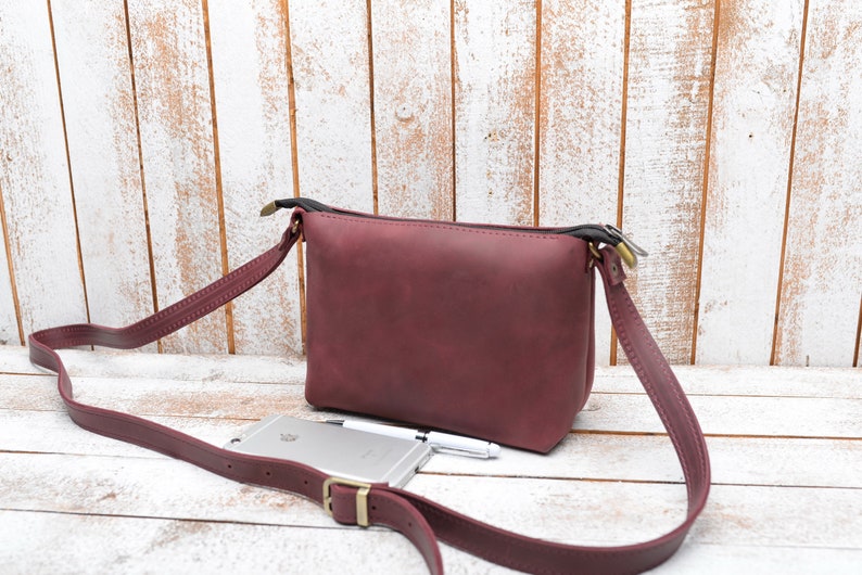 Peut inclure: Un sac &agrave; bandouli&egrave;re en cuir bordeaux avec une longue bandouli&egrave;re r&eacute;glable. Le sac se ferme avec une fermeture &eacute;clair et poss&egrave;de une petite poche sur le devant. Le sac est pos&eacute; sur une surface en bois blanc.