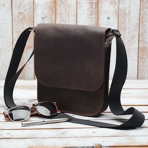 May include: A brown leather messenger bag with a black adjustable strap. The bag is lying on a white wooden surface with a pair of sunglasses and a pen.