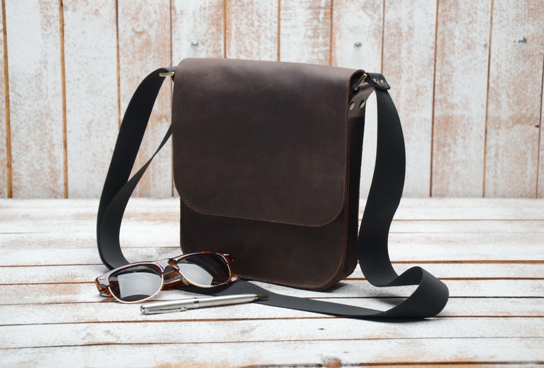 May include: A brown leather messenger bag with a black adjustable strap. The bag is lying on a white wooden surface with a pair of sunglasses and a pen.