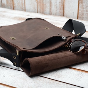 May include: A brown leather messenger bag with a black adjustable strap. The bag has a flap with a small pocket and a zipper closure. Sunglasses are tucked into the bag's open flap.
