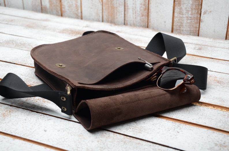 May include: A brown leather messenger bag with a black adjustable strap. The bag has a flap with a small pocket and a zipper closure. Sunglasses are tucked into the bag's open flap.