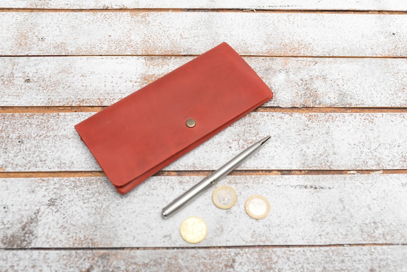 May include: A red leather wallet with a silver pen and three gold coins on a white wooden surface.