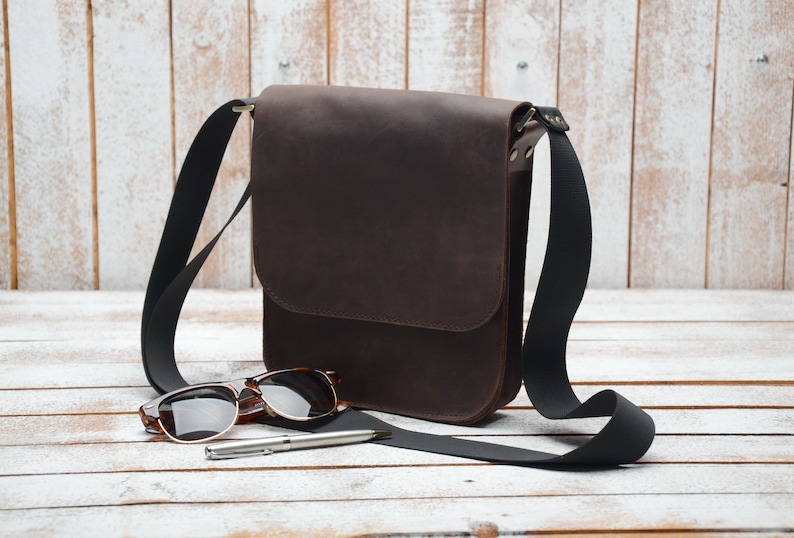 May include: A brown leather messenger bag with a black adjustable strap. The bag is lying on a white wooden surface with a pair of sunglasses and a pen.