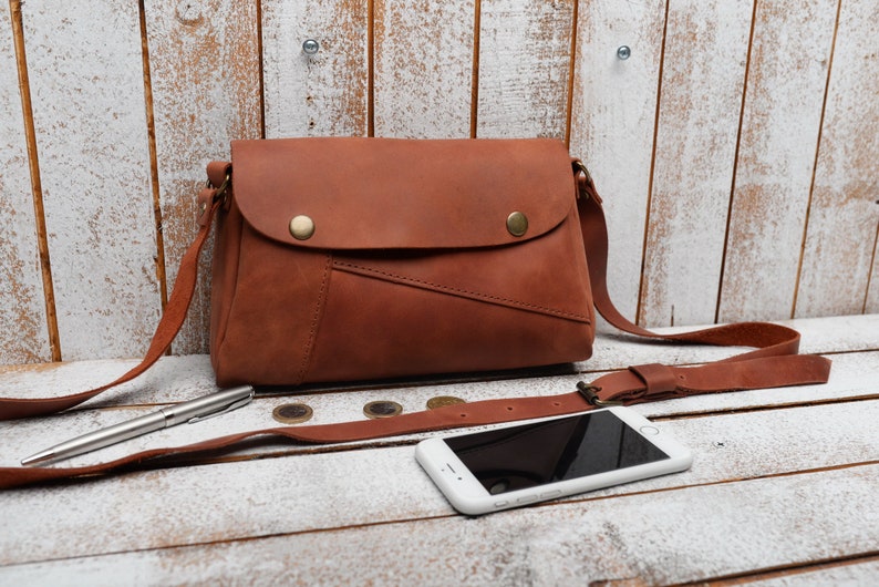 May include: A brown leather crossbody bag with a flap closure and a long adjustable strap. The bag is sitting on a white wooden surface with a pen, coins, and a white smartphone.