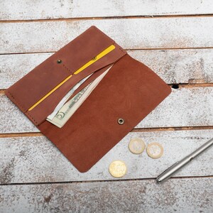 May include: A brown leather wallet with a yellow elastic band, open to reveal cash and a credit card. The wallet is on a white wooden surface with a silver pen and three coins.