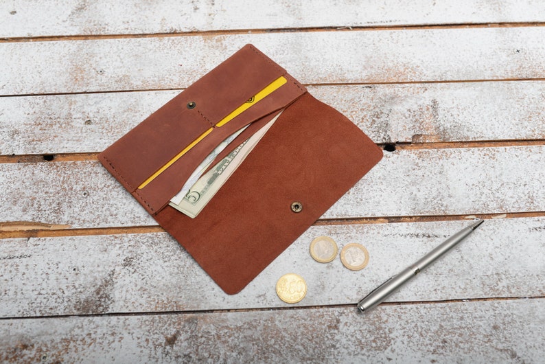 May include: A brown leather wallet with a yellow elastic band, open to reveal cash and a credit card. The wallet is on a white wooden surface with a silver pen and three coins.