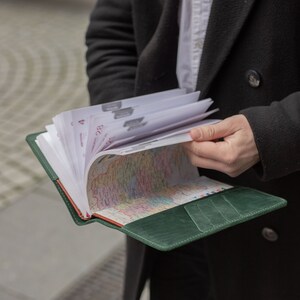 May include: A green leather bound journal with a map inside. The journal is open and the pages are fanned out. The journal is being held by a person wearing a black coat.