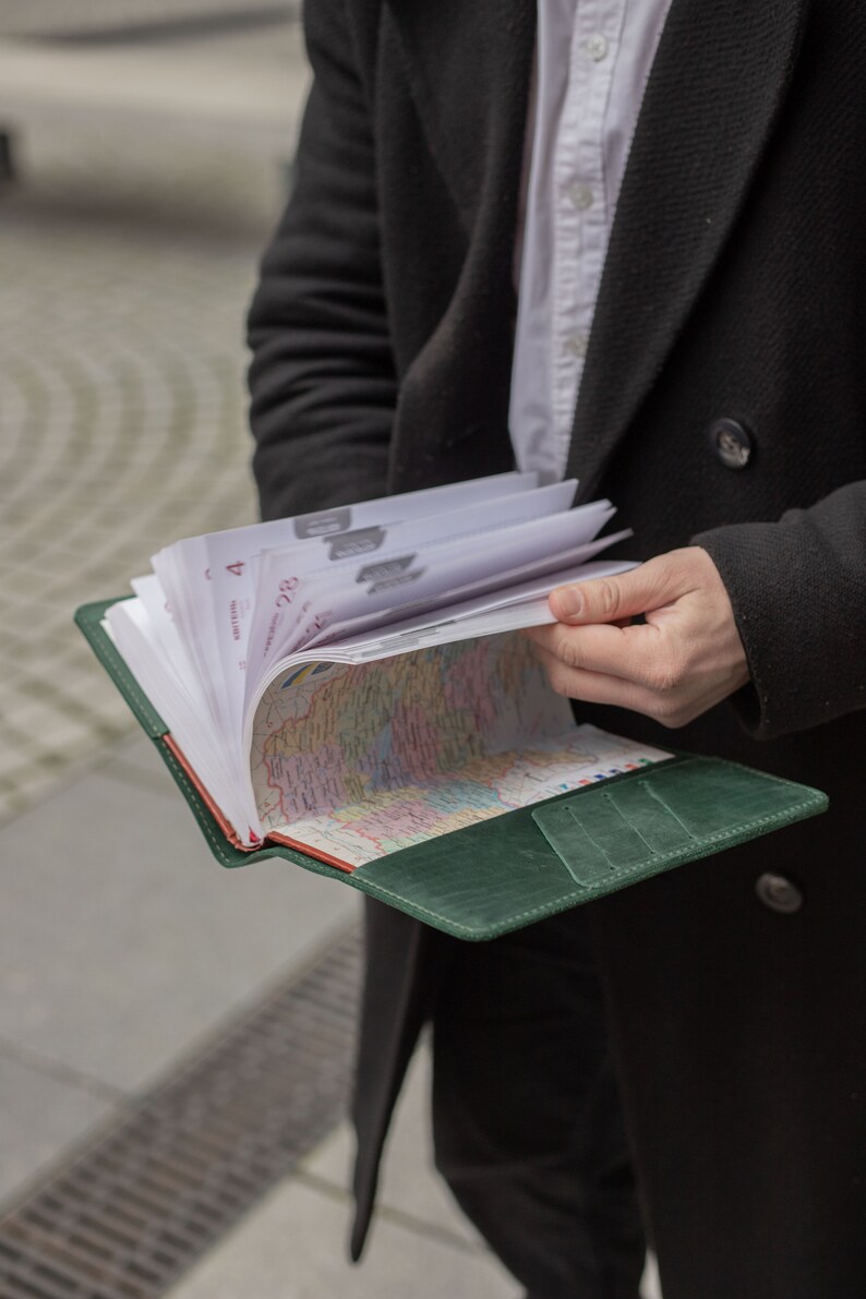 May include: A green leather bound journal with a map inside. The journal is open and the pages are fanned out. The journal is being held by a person wearing a black coat.