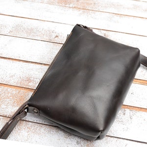 Pu&ograve; includere: Una borsa a tracolla in pelle marrone scuro con chiusura a zip e una lunga tracolla.