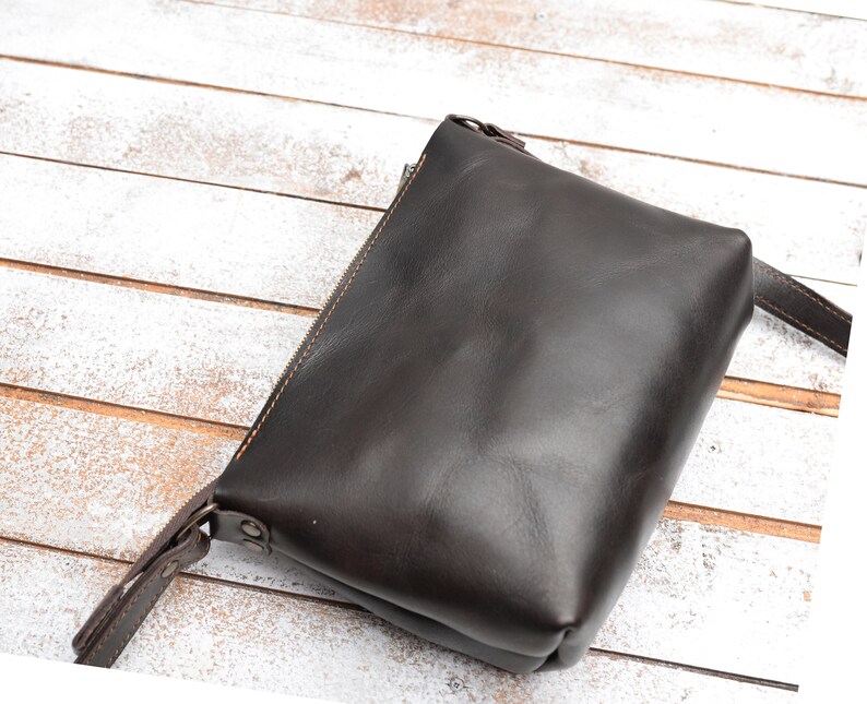 Pu&ograve; includere: Una borsa a tracolla in pelle marrone scuro con chiusura a zip e una lunga tracolla.