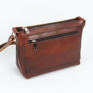 May include: A brown leather crossbody bag with a zippered pocket on the front. The bag has a long adjustable strap.