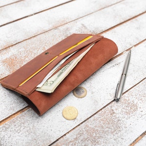 May include: A brown leather wallet with a yellow interior, open to reveal cash and cards. A silver pen and two coins are on a white wooden surface.