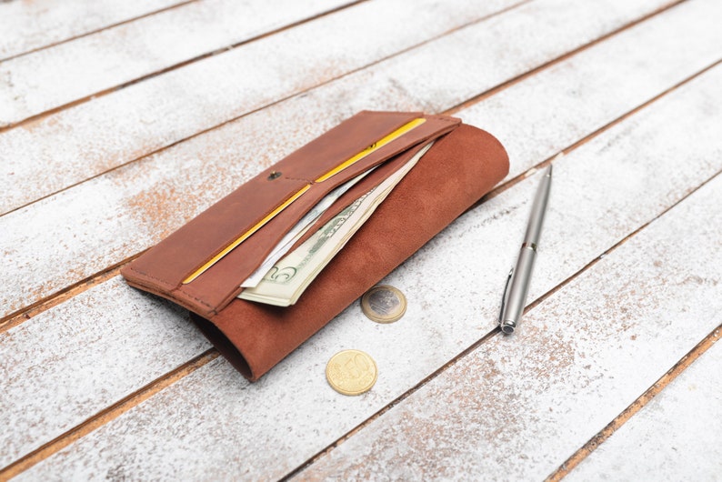 May include: A brown leather wallet with a yellow interior, open to reveal cash and cards. A silver pen and two coins are on a white wooden surface.