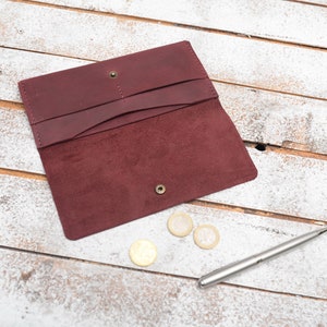 May include: A burgundy leather wallet with a suede interior, open to reveal three compartments. The wallet is lying on a white wooden surface with three gold coins and a silver pen.