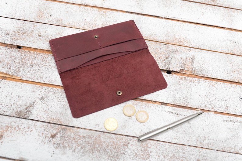 May include: A burgundy leather wallet with a suede interior, open to reveal three compartments. The wallet is lying on a white wooden surface with three gold coins and a silver pen.
