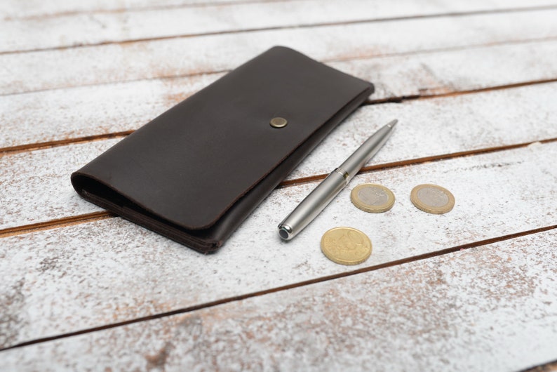 May include: A brown leather wallet with a silver pen and three gold coins on a white wooden surface.