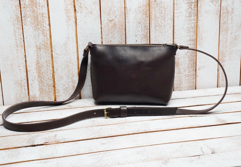 May include: A brown leather crossbody bag with a long adjustable strap and a gold buckle. The bag has a zipper closure and a flap.