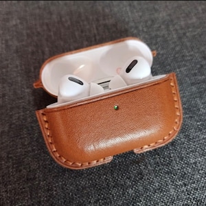 Puede incluir: Una funda de cuero marr&oacute;n para auriculares inal&aacute;mbricos. La funda est&aacute; abierta y los auriculares son visibles en el interior.