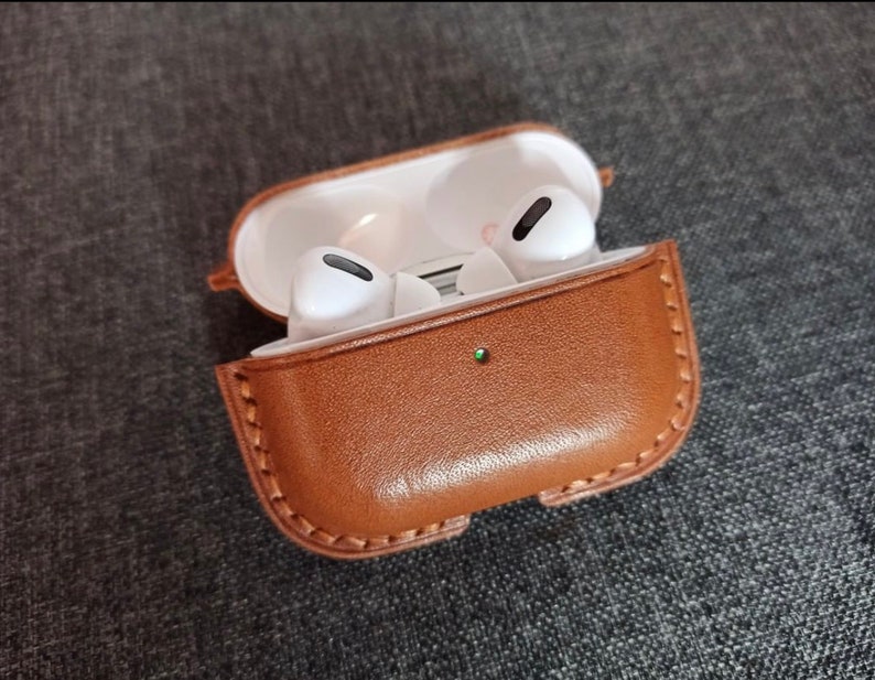 Puede incluir: Una funda de cuero marr&oacute;n para auriculares inal&aacute;mbricos. La funda est&aacute; abierta y los auriculares son visibles en el interior.