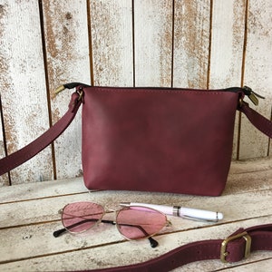 Peut inclure: Un sac &agrave; bandouli&egrave;re en cuir bordeaux avec une fermeture &agrave; glissi&egrave;re et une longue bandouli&egrave;re r&eacute;glable. Le sac est pos&eacute; sur une surface en bois blanc avec une paire de lunettes de soleil roses et un stylo.