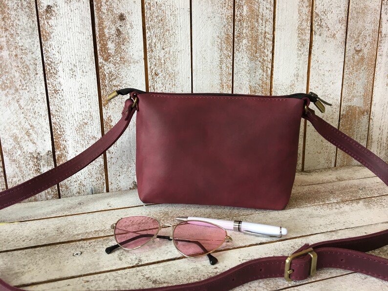 Peut inclure: Un sac &agrave; bandouli&egrave;re en cuir bordeaux avec une fermeture &agrave; glissi&egrave;re et une longue bandouli&egrave;re r&eacute;glable. Le sac est pos&eacute; sur une surface en bois blanc avec une paire de lunettes de soleil roses et un stylo.