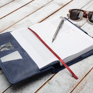 May include: A blue leather-bound journal with a red ribbon bookmark and a silver pen. The journal is open to a blank page with lined paper. There are sunglasses and coins on a white wooden surface.