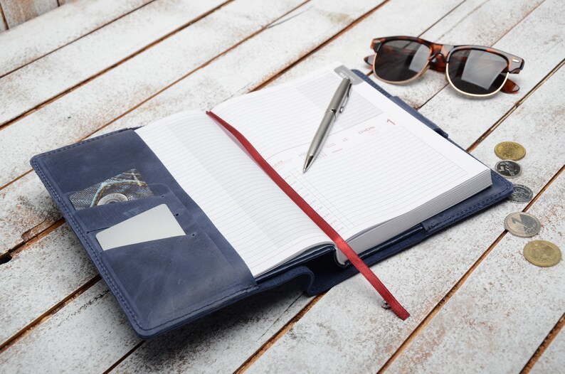 May include: A blue leather-bound journal with a red ribbon bookmark and a silver pen. The journal is open to a blank page with lined paper. There are sunglasses and coins on a white wooden surface.