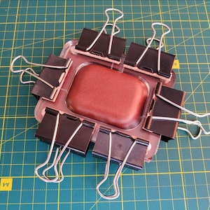 Puede incluir: Un objeto rectangular marr&oacute;n se mantiene en su lugar mediante ocho clips de carpeta negros sobre una estera de corte verde. El objeto est&aacute; rodeado por los clips, que est&aacute;n unidos a los bordes del objeto.