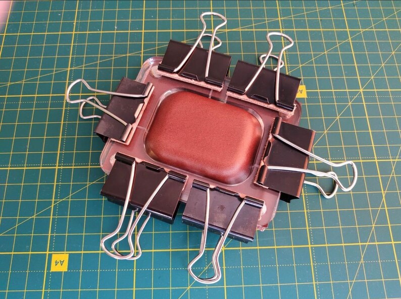 Puede incluir: Un objeto rectangular marr&oacute;n se mantiene en su lugar mediante ocho clips de carpeta negros sobre una estera de corte verde. El objeto est&aacute; rodeado por los clips, que est&aacute;n unidos a los bordes del objeto.