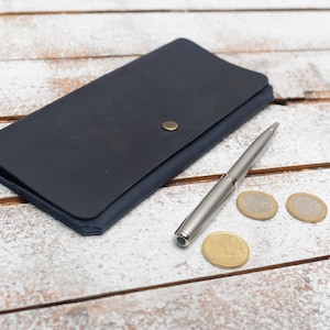 May include: A dark blue leather wallet with a silver pen and three gold coins on a white wooden surface.